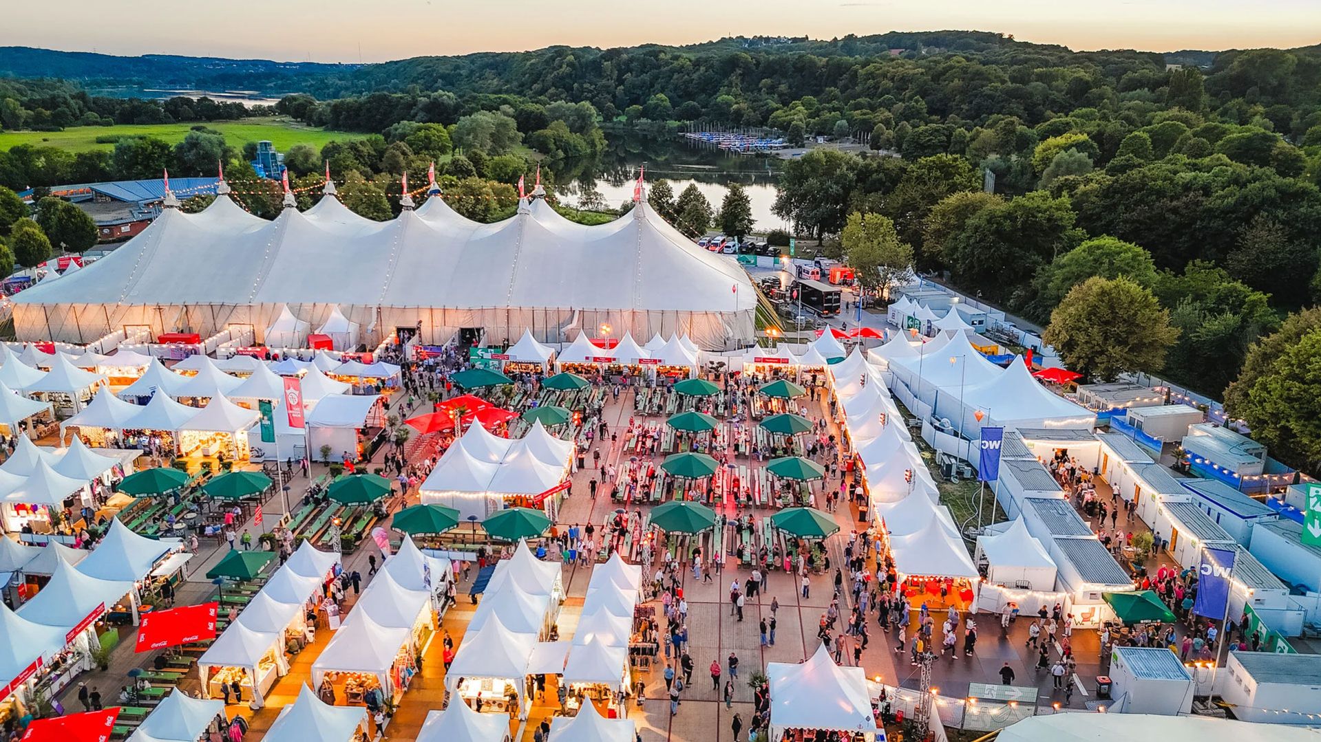 Luftaufnahme eines großen Festzelts mit vielen kleineren weißen Zelten und Menschen auf einem Veranstaltungsgelände neben einem See und Wald